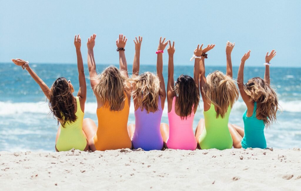 Back view photo of six girls wearing swimsuit sitting on white sand