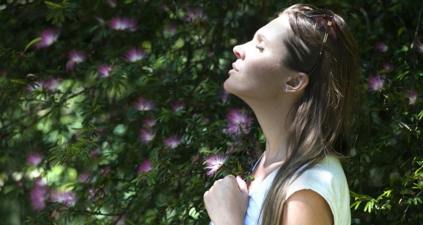 Woman Closing Her Eyes Against Sun Light Standing Near Purple Petaled Flower Plant
