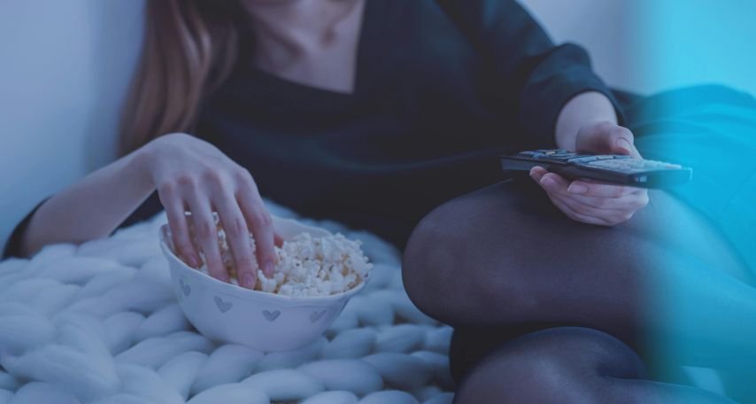 Woman in White Bed Holding Remote Control While Eating Popcorn
