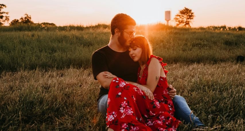 Man in Black Crew Neck T-shirt Sitting on Grass Field Beside Woman in Red Floral Dress
