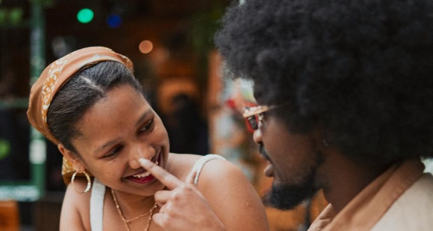 Woman Sitting at a Table with a Man and Pointing at her Nose
