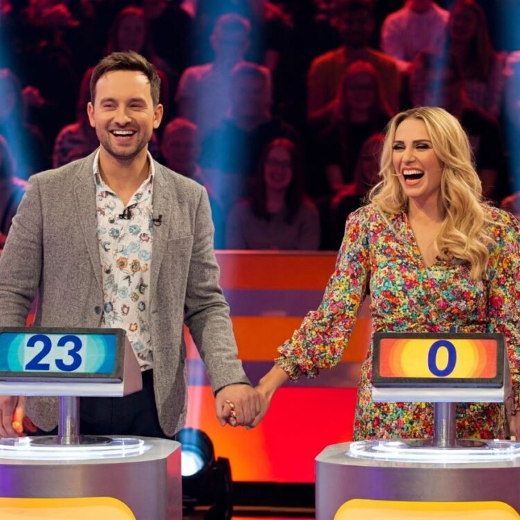 Newlyweds on a brightly lit game show set, smiling and holding hands.