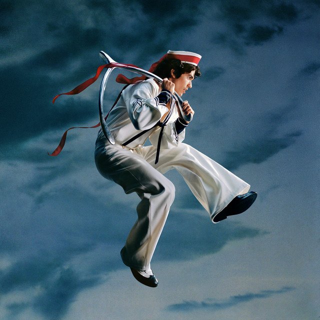 Cover art for Conan Gray’s album “Wishbone,” featuring Conan mid-jump in a white sailor suit with red ribbons flowing, set against a cloudy blue sky.
